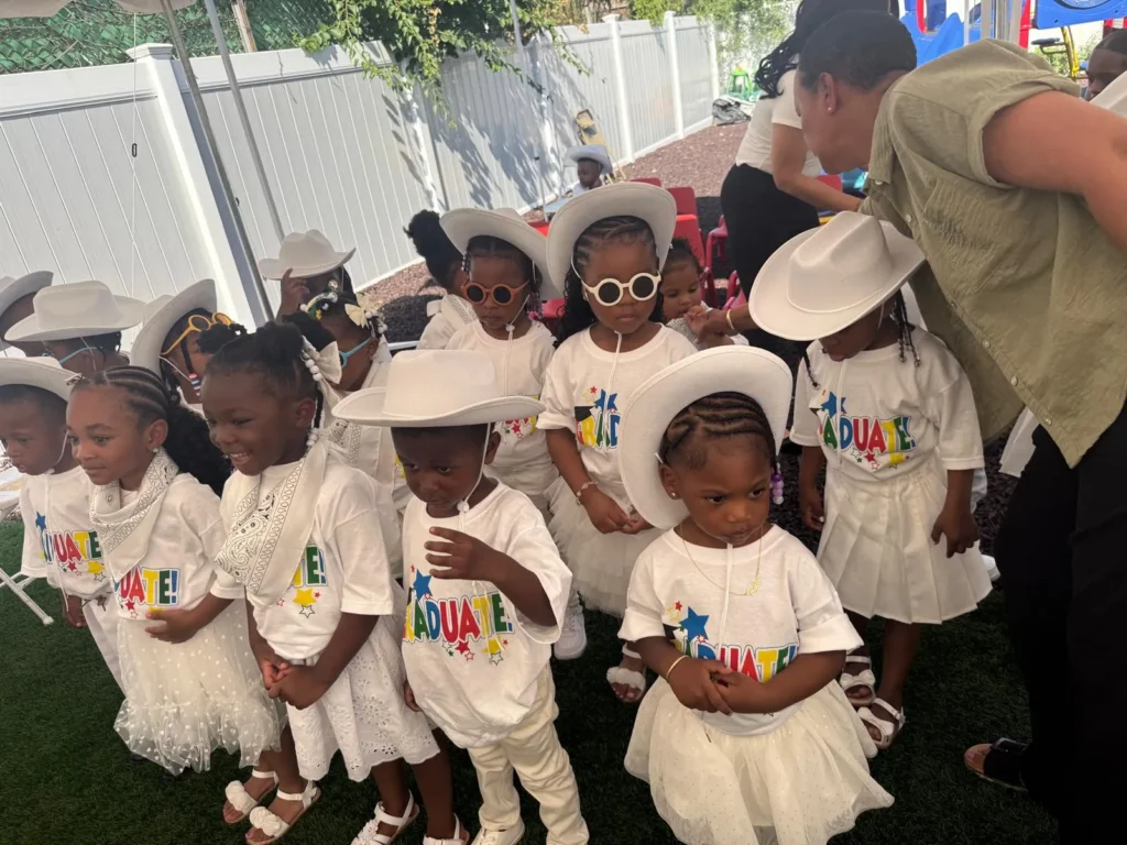 Young children wearing white graduation outfits and cowboy hats at an outdoor school celebration